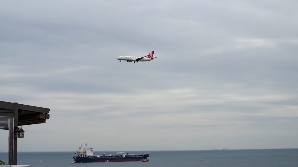 Airplane approaching over the sea with a cargo ship below under a cloudy sky.